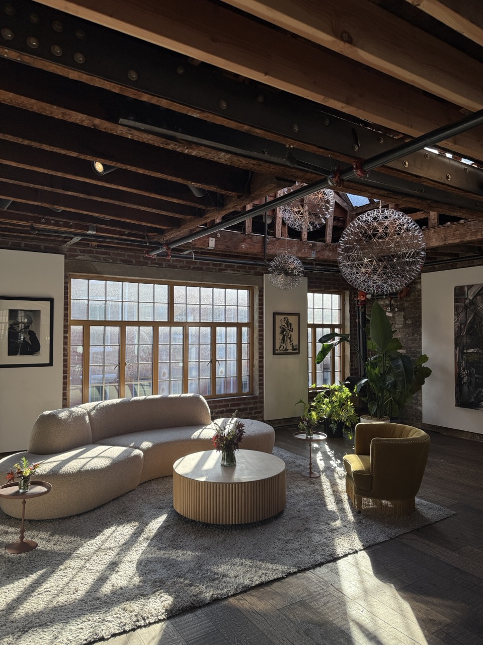 Sunlit industrial loft lounge with exposed brick, wood beams, modern seating, greenery, and hanging sculptural lights.